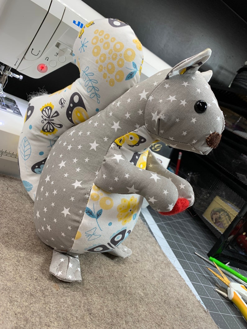 May include: A grey and white fabric squirrel with a yellow and blue butterfly pattern on its back. The squirrel has a red nose and black eyes.