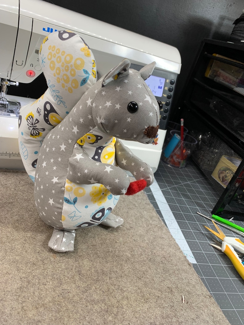May include: A grey and white stuffed squirrel toy with a yellow and white patterned tail. The squirrel is holding a red felt acorn in its paw.