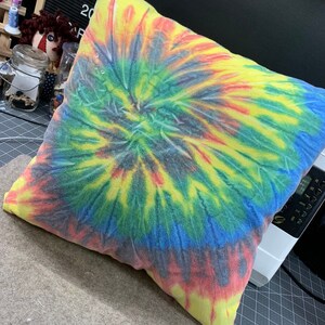 May include: A square pillow with a vibrant tie-dye pattern in a spiral design. The colors include yellow, red, blue, and green. The pillow is on a gray surface.