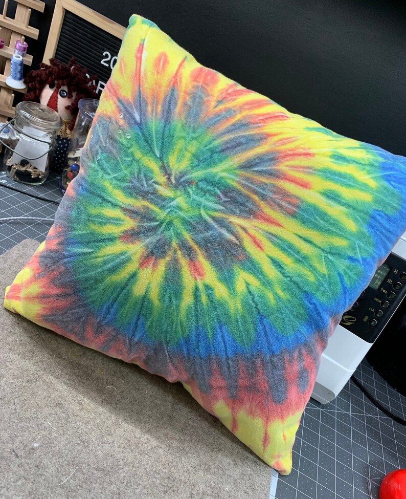 May include: A square pillow with a vibrant tie-dye pattern in a spiral design. The colors include yellow, red, blue, and green. The pillow is on a gray surface.