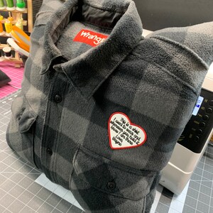 May include: A gray and black plaid flannel shirt with a red heart-shaped patch that says "This is a shirt I used to wear and whenever you hold it know I am there Love Wayne". The shirt has a Wrangler label on the collar.
