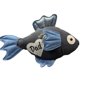 May include: A plush fish toy in shades of gray and blue, with a heart-shaped patch that reads "Dad." The fish has a large eye and fins made of denim-like fabric. The word "AURO" is visible on the fish.
