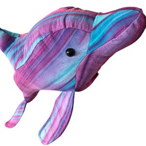 May include: A handmade plush whale toy in shades of purple, blue, and pink. The whale has a black button eye and stitched details. The fabric has a swirled pattern. The fins and tail are visible.
