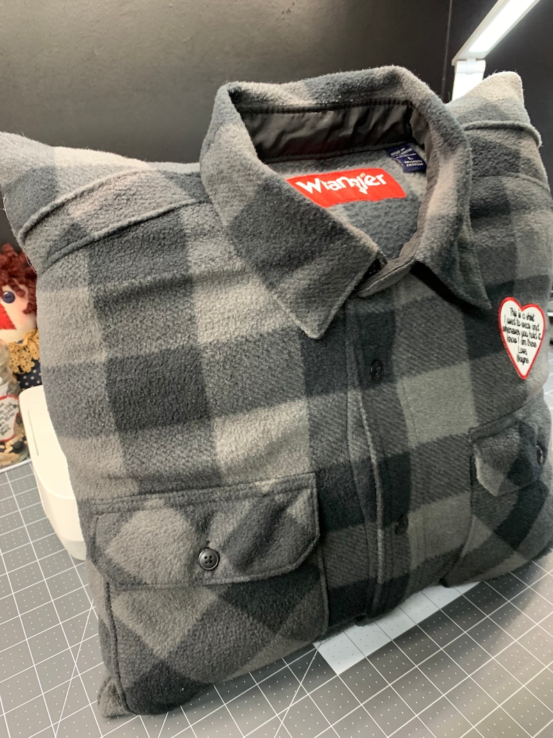 May include: A gray and black plaid flannel shirt pillow with a red "Wrangler" label and a heart-shaped patch with a message.