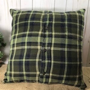 May include: Square pillow with a green, navy, and cream plaid pattern. The pillow cover appears to be made from a button-down shirt, with the buttons and pocket visible. The pillow is on a burlap surface.