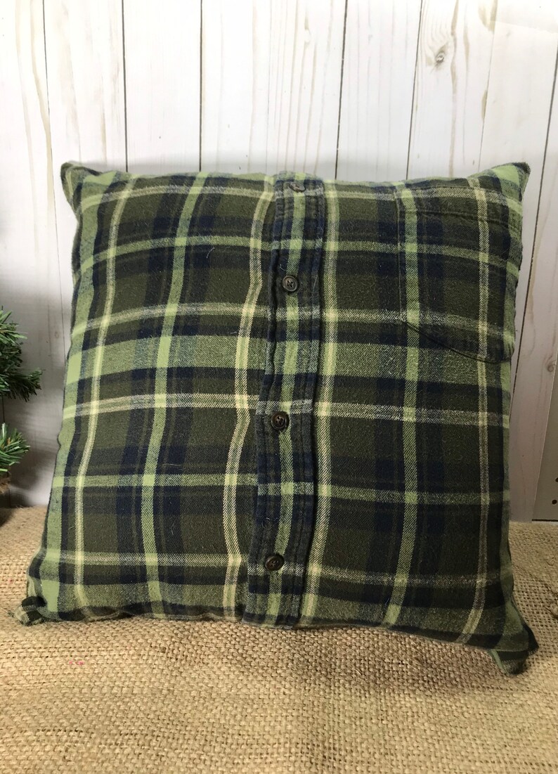 May include: Square pillow with a green, navy, and cream plaid pattern. The pillow cover appears to be made from a button-down shirt, with the buttons and pocket visible. The pillow is on a burlap surface.