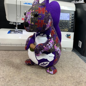 May include: A purple and white stuffed squirrel with a jigsaw puzzle pattern. The squirrel is holding a small brown acorn in its paws.