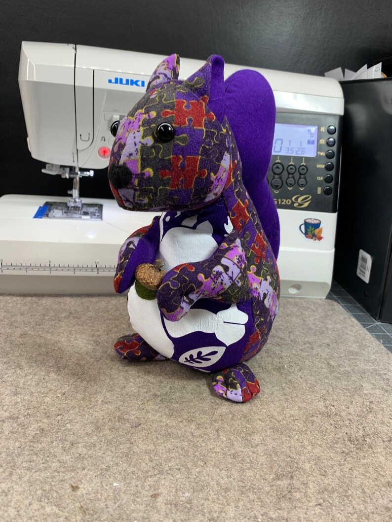 May include: A purple and white stuffed squirrel with a jigsaw puzzle pattern. The squirrel is holding a small brown acorn in its paws.