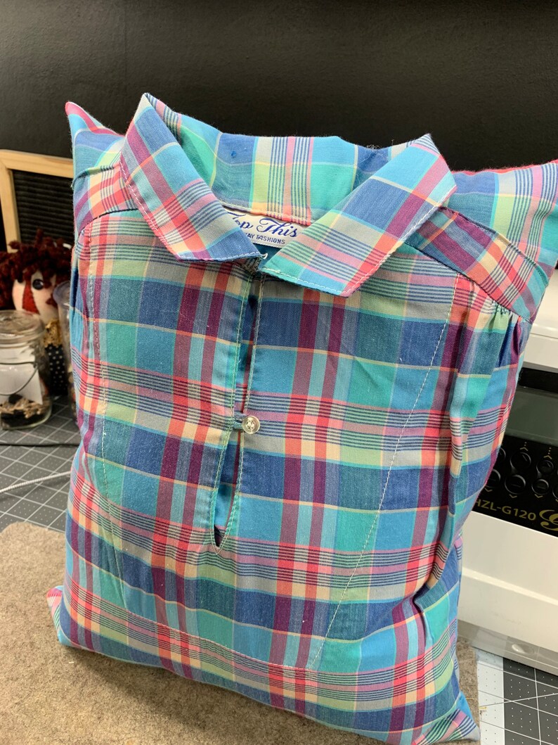May include: A blue, pink, and green plaid pillow with a button closure. The pillow is made from a repurposed shirt with the text "Top This" on the collar.