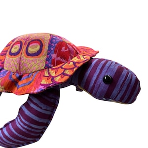 May include: A colorful stuffed turtle with a patterned shell in shades of red, orange, and purple. The turtle's head and limbs are purple with light purple stripes. The turtle has a black button eye.
