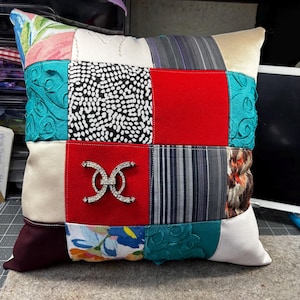 May include: Square patchwork pillow with a variety of fabric patterns and colors. The pillow features a red square with a silver-colored decorative embellishment. Other fabrics include floral, polka dot, and striped patterns. The pillow is a decorative home accent.