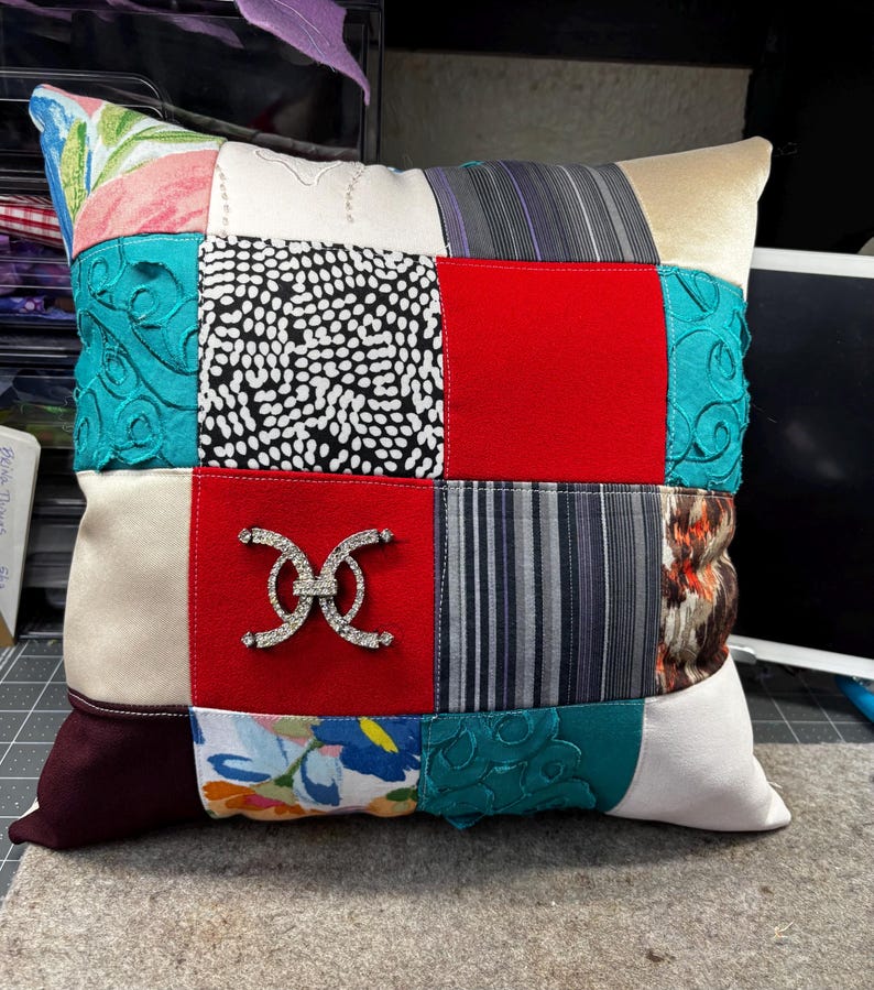 May include: Square patchwork pillow with a variety of fabric patterns and colors. The pillow features a red square with a silver-colored decorative embellishment. Other fabrics include floral, polka dot, and striped patterns. The pillow is a decorative home accent.