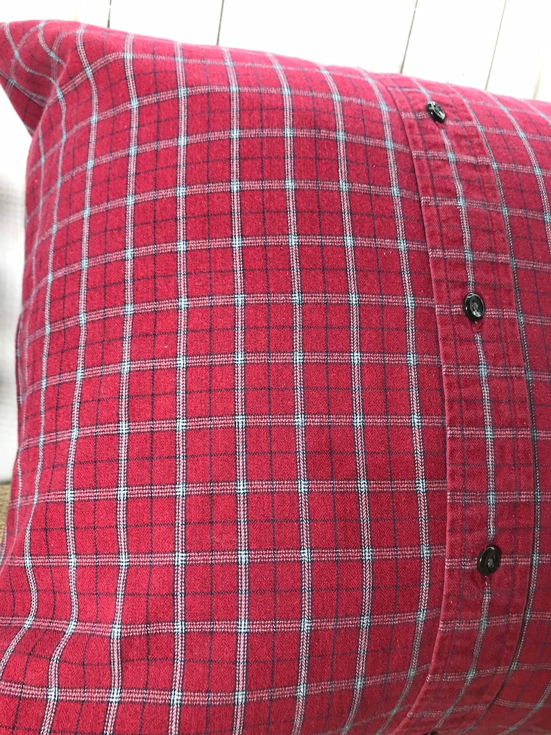 May include: A square pillow with a red and black plaid pattern. The pillow is made from a repurposed button-down shirt, with black buttons visible on the side. The pillow is a decorative home accent.