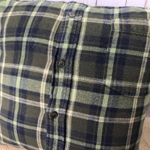 May include: Square pillow with a green, navy, and white plaid pattern. The pillow is made from a button-down shirt, with the buttons and pocket visible. The pillow has a rustic, farmhouse style.