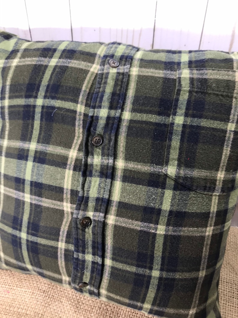 May include: Square pillow with a green, navy, and white plaid pattern. The pillow is made from a button-down shirt, with the buttons and pocket visible. The pillow has a rustic, farmhouse style.