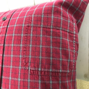 May include: A square pillow made from a red plaid shirt. The pillow features a button-down front, a pocket, and a classic plaid pattern in red, black, and light blue. The pillow is a decorative home accent.