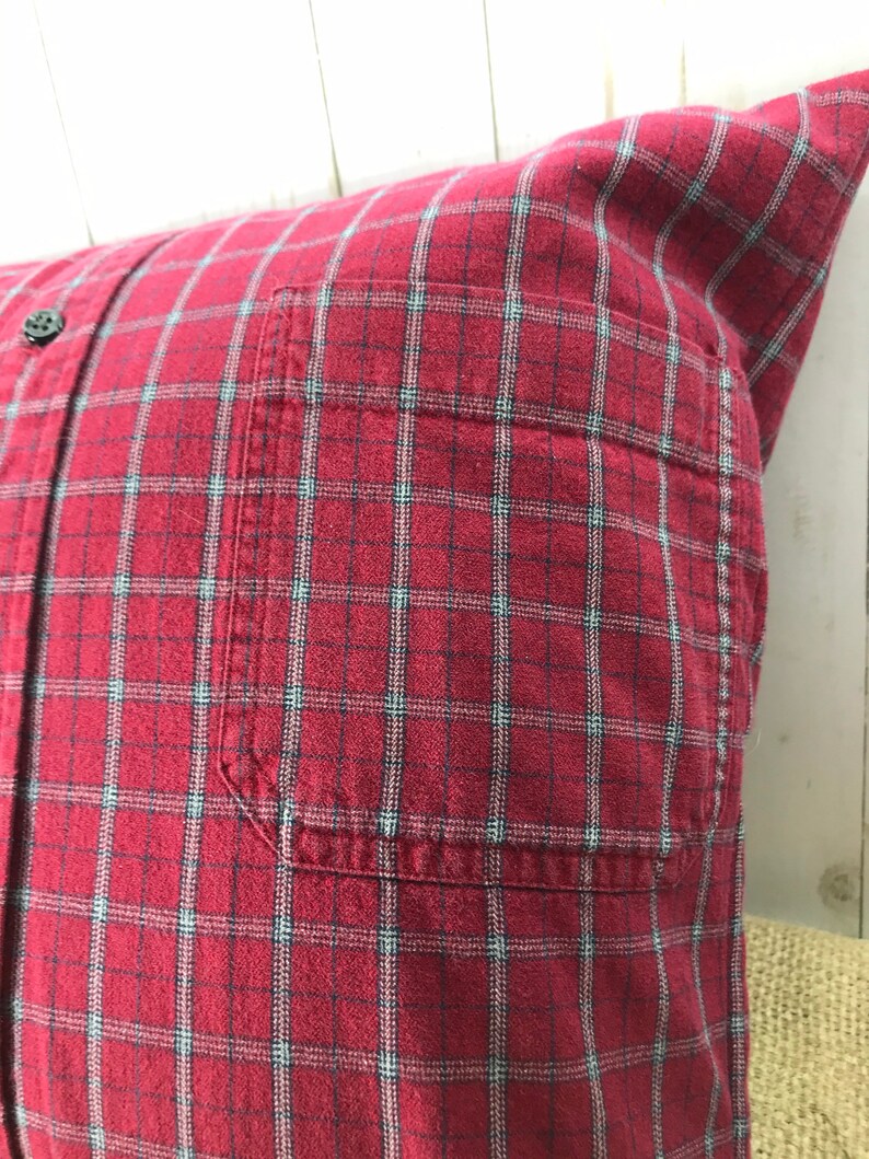 May include: A square pillow made from a red plaid shirt. The pillow features a button-down front, a pocket, and a classic plaid pattern in red, black, and light blue. The pillow is a decorative home accent.