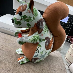 May include: A brown and green fabric squirrel with a bushy tail. The squirrel is holding a red acorn in its paws. The fabric has a pattern of pine trees, berries, and squirrels.