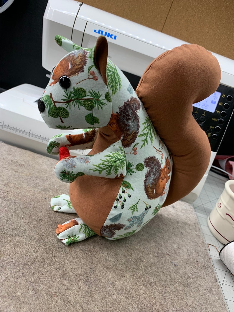 May include: A brown and green fabric squirrel with a bushy tail. The squirrel is holding a red acorn in its paws. The fabric has a pattern of pine trees, berries, and squirrels.