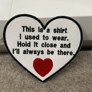 May include: White heart-shaped embroidered patch with black stitching and the text "This is a shirt I used to wear. Hold it close and I'll always be there."  A red embroidered heart is in the center of the patch.