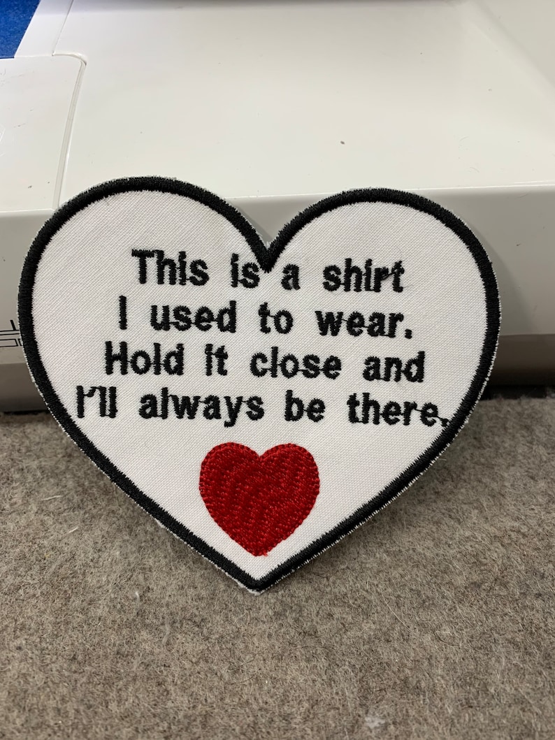 May include: White heart-shaped embroidered patch with black stitching and the text "This is a shirt I used to wear. Hold it close and I'll always be there."  A red embroidered heart is in the center of the patch.