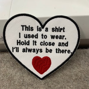 May include: A heart-shaped embroidered patch with a white background and a black border. The patch features black embroidered text that reads: This is a shirt I used to wear. Hold it close and I'll always be there. A small red heart is below the text.