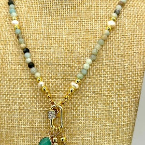 May include: A beaded necklace with a gold chain and multiple charms. The necklace features a key, a cross, and a green stone pendant. The beads are a mix of colors, including green, white, and black. The necklace is displayed on a tan fabric.