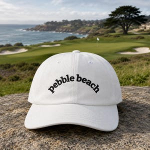 Op de afbeelding: Een witte baseballcap met de tekst "pebble beach" in het zwart geborduurd. De cap ligt op een rots, met een golfbaan en de zee op de achtergrond.