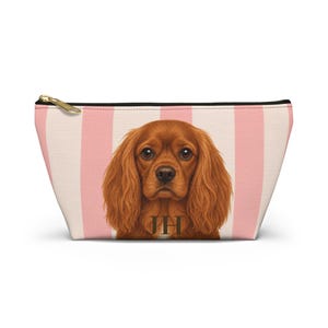 Può includere: Una pochette trapezoidale con un motivo a righe verticali rosa chiaro e bianco. Al centro è presente un'illustrazione dettagliata di un Cavalier King Charles Spaniel marrone con le lettere del monogramma JH in marrone scuro.