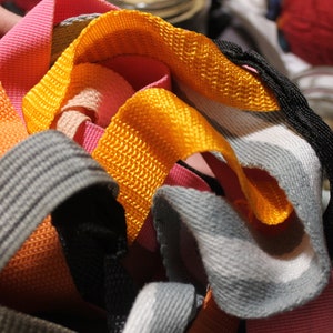 May include: A close-up of a pile of colorful woven straps in orange, pink, gray, and yellow. The straps are made of a woven fabric and are likely used for crafting or DIY projects.