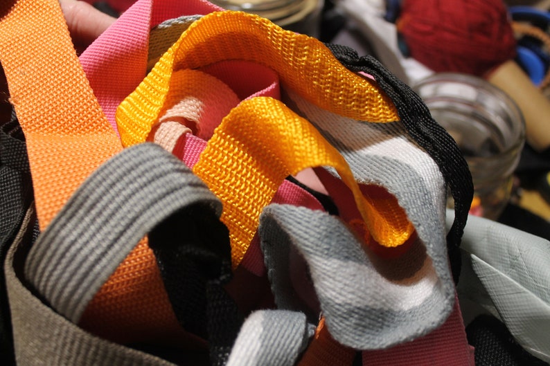 May include: A close-up of a pile of colorful woven straps in orange, pink, gray, and yellow. The straps are made of a woven fabric and are likely used for crafting or DIY projects.