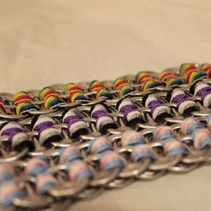 May include: Close-up of several handmade chainmail-style bracelets. Each bracelet is made from silver-colored metal rings and features colorful ribbon accents. The ribbons are in rainbow, purple, and light blue and pink patterns.
