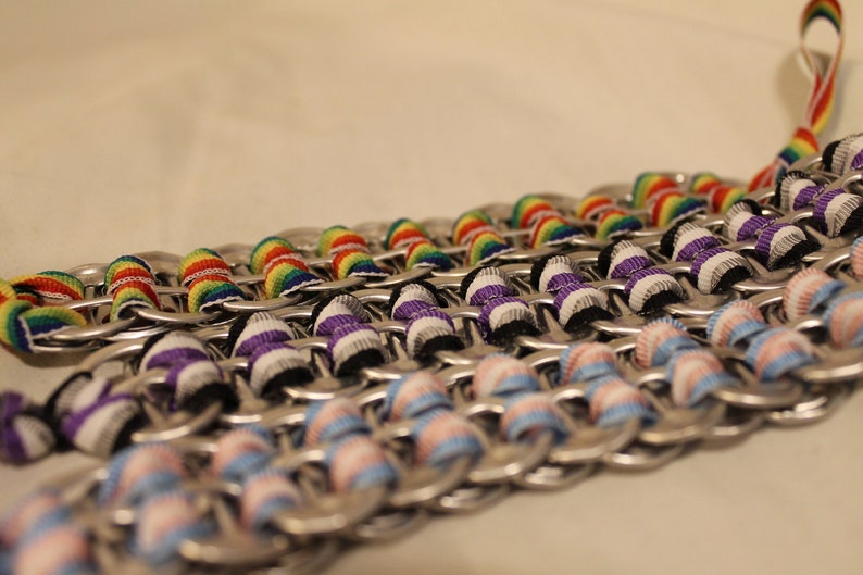 May include: Close-up of several handmade chainmail-style bracelets. Each bracelet is made from silver-colored metal rings and features colorful ribbon accents. The ribbons are in rainbow, purple, and light blue and pink patterns.