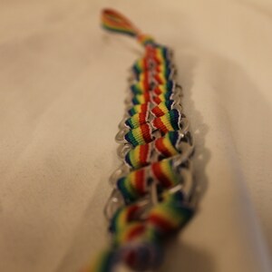 May include: A handmade bracelet featuring a woven design with rainbow-colored ribbons and silver-colored metal tabs. The bracelet is a vibrant accessory, showcasing a spectrum of colors.