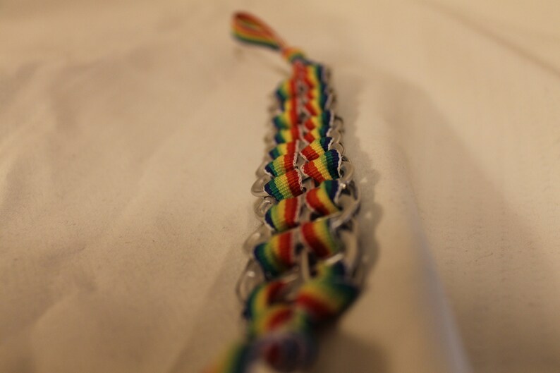May include: A handmade bracelet featuring a woven design with rainbow-colored ribbons and silver-colored metal tabs. The bracelet is a vibrant accessory, showcasing a spectrum of colors.