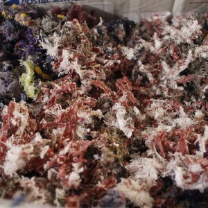 May include: Close-up of a box filled with colorful shredded fabric. The fabric is a mix of red, white, blue, purple, and yellow. The shredded material appears to be a mix of different textures and weights.