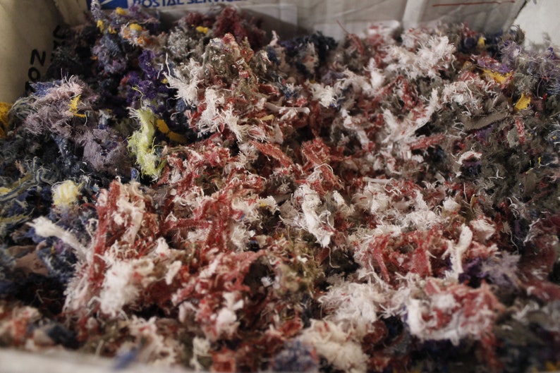 May include: Close-up of a box filled with colorful shredded fabric. The fabric is a mix of red, white, blue, purple, and yellow. The shredded material appears to be a mix of different textures and weights.