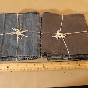 May include: Two stacks of denim fabric squares, tied with twine, are arranged side-by-side on a brown surface. A wooden ruler with inch markings is placed below the stacks.