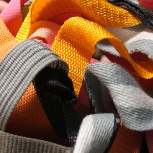 May include: A close-up of a pile of colorful woven straps in orange, gray, black, and pink. The straps are made of a textured fabric and are likely used for crafting or sewing projects.
