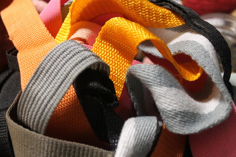 May include: A close-up of a pile of colorful woven straps in orange, gray, black, and pink. The straps are made of a textured fabric and are likely used for crafting or sewing projects.