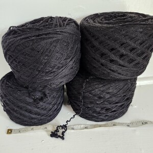 May include: Four round balls of black yarn arranged in a square formation. A white measuring tape is positioned horizontally in front of the yarn, showing a length of approximately 10 inches across the base of the four yarn balls.