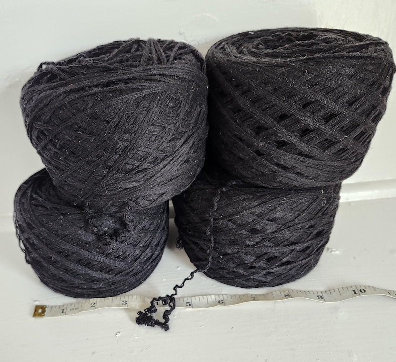 May include: Four round balls of black yarn arranged in a square formation. A white measuring tape is positioned horizontally in front of the yarn, showing a length of approximately 10 inches across the base of the four yarn balls.