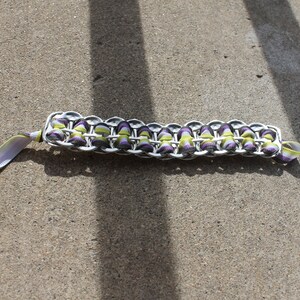 May include: A handmade bracelet crafted from recycled aluminum can tabs. The bracelet features a woven design with purple and green accents, and a matching ribbon for closure. The bracelet is shown on a concrete surface.