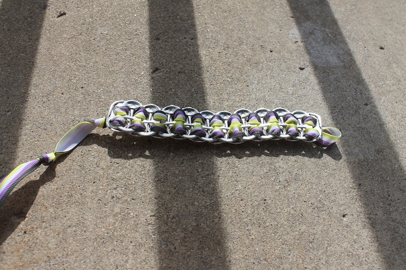 May include: A handmade bracelet crafted from recycled aluminum can tabs. The bracelet features a woven design with purple and green accents, and a matching ribbon for closure. The bracelet is shown on a concrete surface.