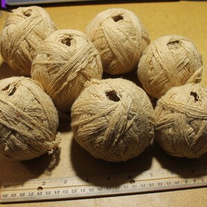 May include: Seven round balls of light tan, textured twine arranged in a cluster. A wooden ruler is positioned at the bottom, showing the balls range from approximately 2 to 3 inches in diameter. The twine has a fibrous, natural appearance.