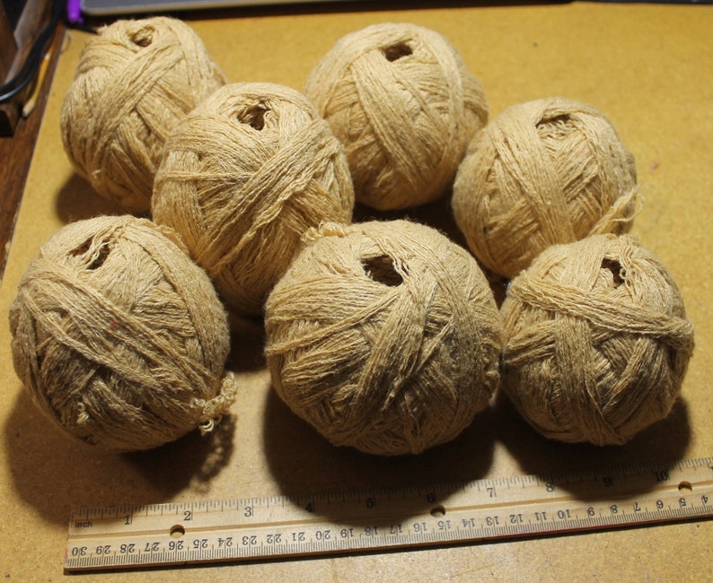 May include: Seven round balls of light tan, textured twine arranged in a cluster. A wooden ruler is positioned at the bottom, showing the balls range from approximately 2 to 3 inches in diameter. The twine has a fibrous, natural appearance.