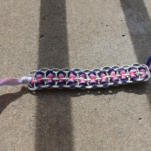 May include: A handmade bracelet crafted from recycled aluminum can tabs, featuring a woven design with pink and purple ribbon accents. The bracelet has a silver metallic finish and includes ribbon ties for closure.
