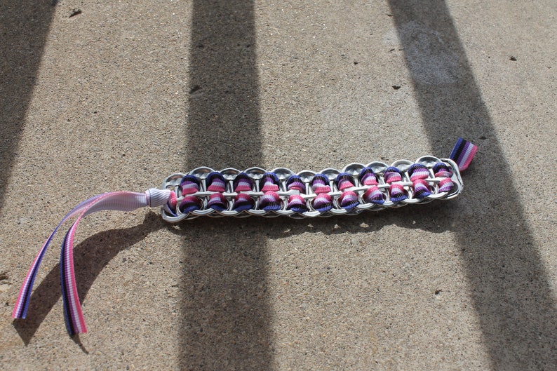 May include: A handmade bracelet crafted from recycled aluminum can tabs, featuring a woven design with pink and purple ribbon accents. The bracelet has a silver metallic finish and includes ribbon ties for closure.