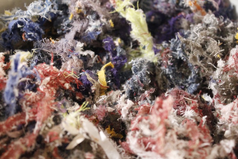 May include: Close-up of a pile of shredded fabric scraps in various colors. The scraps include shades of red, blue, purple, yellow, and gray. The texture is rough and fibrous, with visible threads and frayed edges. The image suggests a craft or textile project.