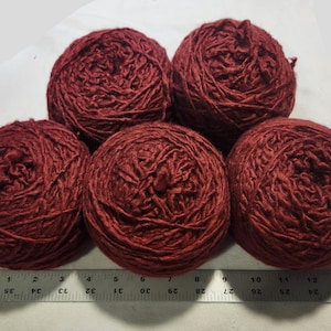 May include: Five round balls of deep red yarn arranged in a cluster. A ruler is positioned along the bottom edge, showing the width of the yarn balls spans approximately 13 inches across. The yarn has a textured, slightly twisted appearance.